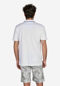 Weißes Polohemd mit Kragen, das mit einem marineblauen Streifen verziert ist, kombiniert mit hellgrauen Shorts, die ein Blattmuster zeigen.
