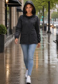 Femme marchant sur un trottoir de ville mouillé avec un imperméable noir à capuche, un jean bleu clair et des baskets blanches par un jour de pluie.