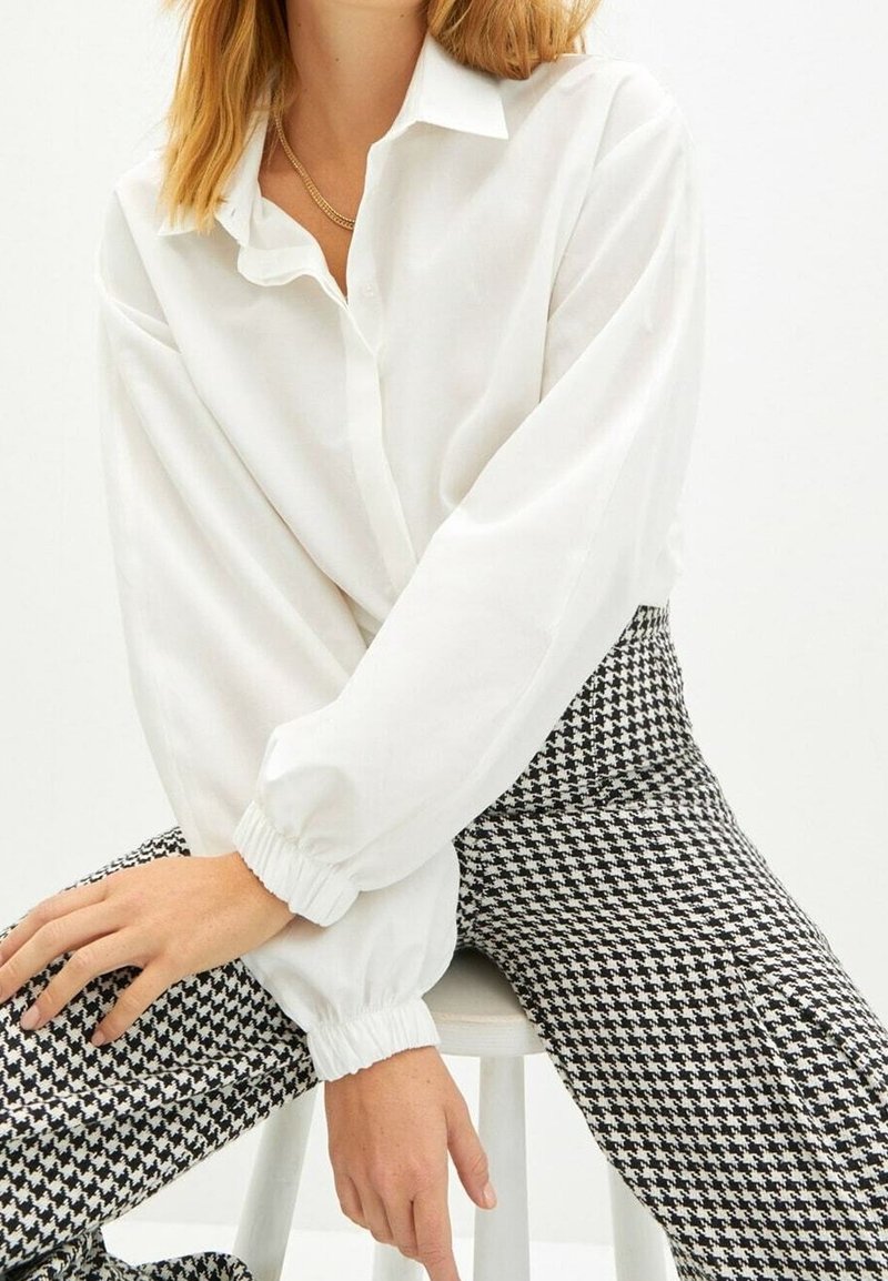 Femme portant un chemisier blanc à manches longues et un pantalon pied-de-poule noir et blanc assise sur un tabouret blanc.