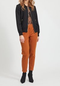 Vrouw in een zwarte bomberjack, met een bruin patroon top, roestkleurige broek en zwarte enkellaarsjes, staand tegen een effen witte achtergrond.