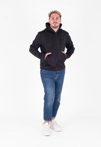 Felpa nera con zip e cappuccio regolabile, tasche anteriori, abbinata a jeans blu e sneakers bianche. Tessuto morbido, design casual.