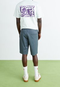 T-shirt vert clair avec un motif violet au dos, associé à un short gris. Chaussures légères et chaussettes blanches. Fond vert.