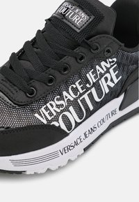Černé tenisky s prodyšným svrškem a bílým logem "Versace Jeans Couture", kulaté tkaničky a texturovaná gumová podrážka.
