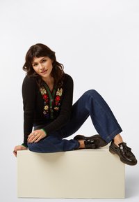 Zwarte trui met bloemenborduurwerk, blauwe wijde denim broek en bruine leren schoenen met kwastjes, zittend op een witte sokkel.