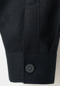 Detailní záběr manžety košile z černé látky s jedním černým knoflíkem a viditelnými švy.