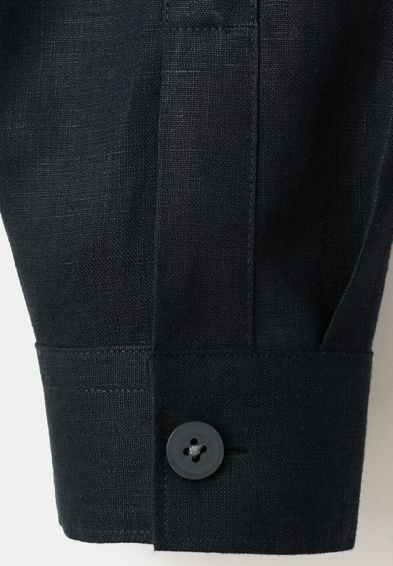 Gros plan sur le poignet d'une chemise en tissu noir avec un seul bouton noir et des détails de couture visibles.