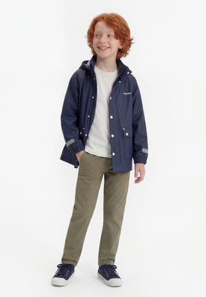 Giovane ragazzo con capelli rossi che indossa un giubbotto antipioggia blu navy, una maglietta bianca, pantaloni oliva e scarpe da ginnastica blu navy, in piedi con una mano in tasca e sorridente.