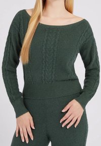 Femme portant un pull en maille torsadée vert foncé à large encolure et un pantalon en tricot assorti, debout les mains près des hanches.