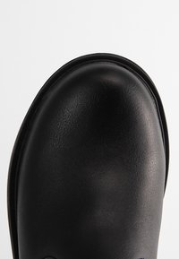 Gros plan sur l'avant d'une chaussure en cuir noir lisse, avec une forme arrondie et un bord cousu, sur un fond clair.