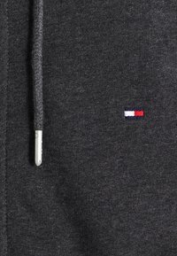 Hoodie gris avec un cordon de serrage, arborant un petit logo brodé en rouge, blanc et bleu sur la poitrine, fabriqué en tissu doux.
