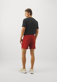 Mann trägt ein schwarzes Kurzarmhemd, rote Sportshorts und hellfarbene Turnschuhe, steht und schaut zur Seite vor einem weißen Hintergrund.