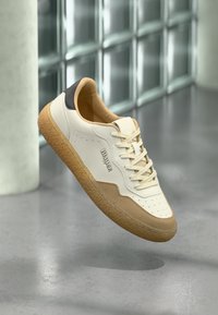 White leather sneaker with a tan suede accent, cream laces, and a gum rubber sole. Features perforations and a black heel tab.