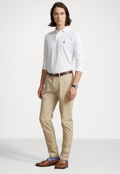 Witte lange mouw polo met een klein logo, beige chino's, bruine loafers en blauwe sokken, gedragen met een bruine riem en een horloge.