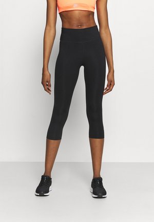 Frau trägt schwarze kurze Leggings, neonorangen Sport-BH und schwarze Laufschuhe, steht vor einem einfarbigen hellen Hintergrund.