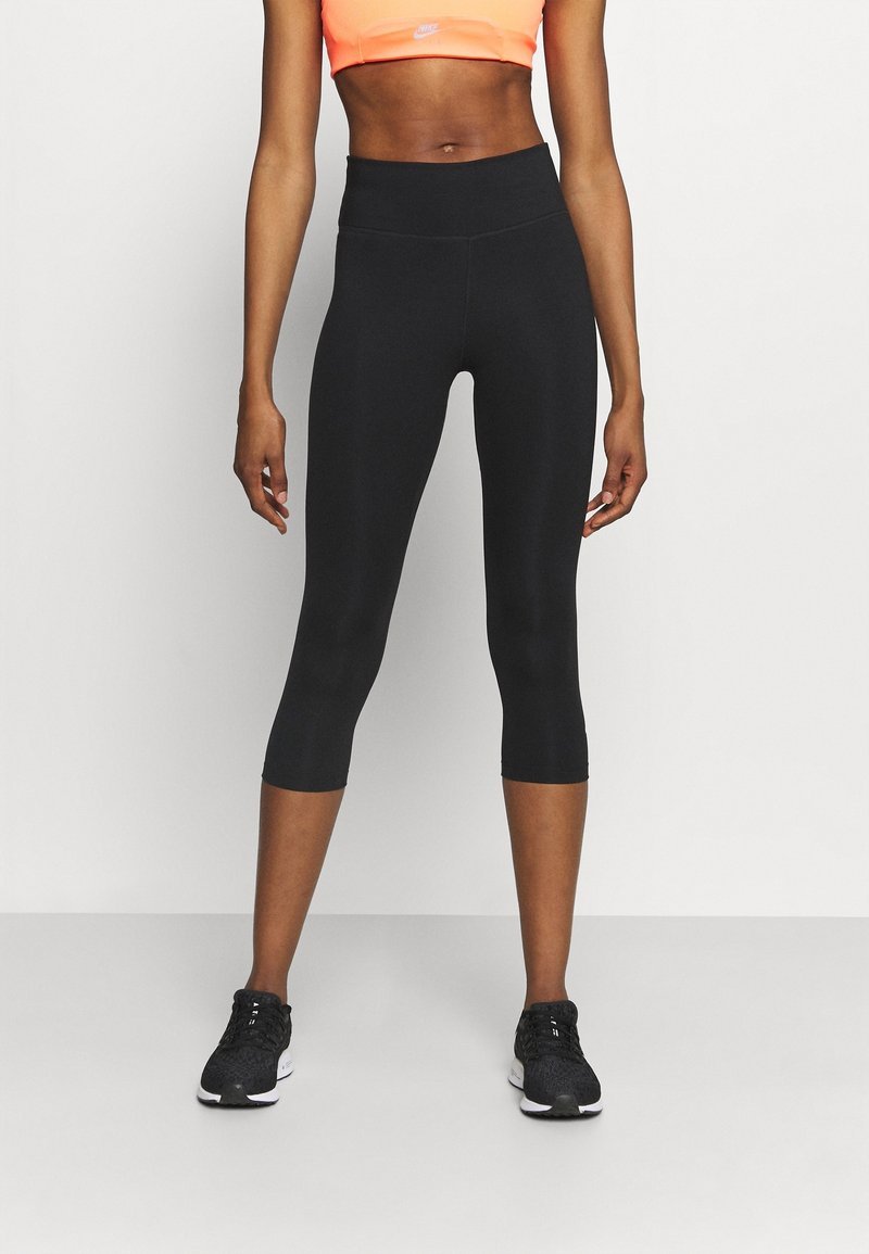 Femme portant un legging court noir, un soutien-gorge de sport orange néon et des chaussures de course noires, debout devant un fond clair uni.