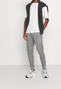 Man som bär en vit t-shirt, en mörkgrå tröja över axlarna, grå joggers med gult logotyp och vita sneakers med svarta och orange detaljer.