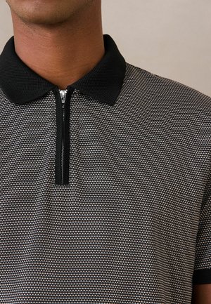 Közeli kép egy személyről, aki texturált fekete-fehér galléros pólót visel fekete gallérral és félig cipzáras lezárással.