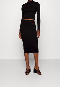 Mujer vestida con un top de manga larga ajustado negro y una falda midi a juego, usando sandalias de tacón alto negras y de punta abierta sobre un fondo sencillo.