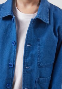 Personne portant un t-shirt blanc à col rond sous une veste bleue boutonnée avec un col et une poche avant visible.