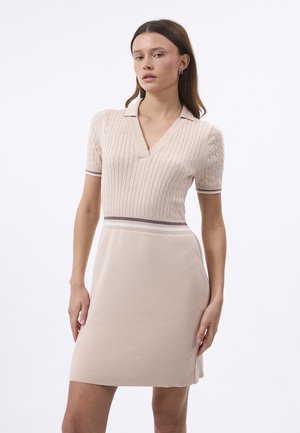 Kvinde iført en beige strikket kjole med korte ærmer, en krave, kabellstrikmønster øverst og stribede detaljer ved taljen og ærmekanterne.