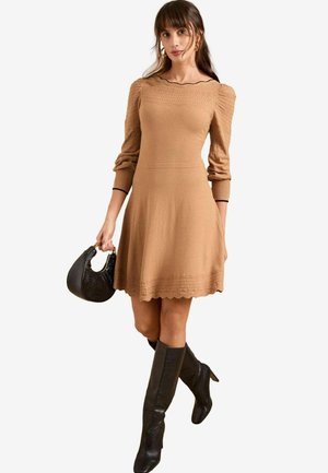 Robe en tricot beige avec un corsage texturé et un décolleté en vagues. Manches longues et bouffantes avec des poignets côtelés. Associée à des bottes noires hautes et un sac à main.