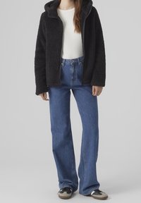 Kvinna klädd i svart fleece zip-up hoodie, vit ribbad skjorta, blå högmidjade jeans och svart-vita sneakers som står mot en grå bakgrund.