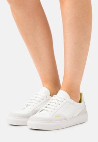 Witte sneakers met lichtgrijze accenten, gestructureerde rubberen zool, vetersluiting en een geel logo aan de zijkant.