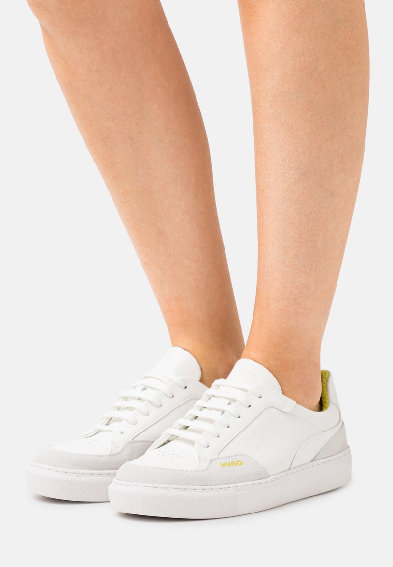 Witte sneakers met lichtgrijze accenten, gestructureerde rubberen zool, vetersluiting en een geel logo aan de zijkant.