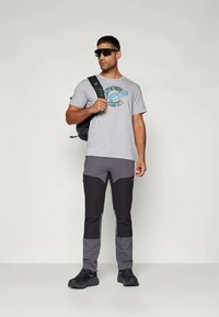 Šedé tričko s barevným logem "Sportswear", tmavě šedé a černé sportovní kalhoty, brýle a černý batoh. Stojící postoj.