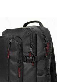 Black backpack made of durable fabric, featuring multiple zippered pockets with red accents, a top handle, and adjustable straps.