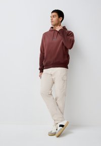 Sweat à capuche marron avec cordon, pantalon cargo crème et baskets avec corps blanc et bande noire. Tissu lisse et coupe décontractée.