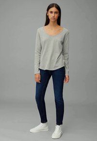 Mulher de pé contra um fundo cinza, vestindo uma blusa de manga longa cinza claro, jeans skinny azul escuro e ténis brancos de cano alto.