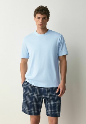 Fiatal férfi göndör hajjal, világoskék rövid ujjú pólót és sötétkék kockás rövidnadrágot visel, egyszínű háttér előtt áll.