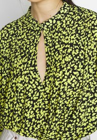 Černá blůza s jasně zeleným květinovým vzorem, výstřihem ve tvaru klíčové dírky, pliseerová konstrukce, hladká textura a nabírané detaily na ramenou.