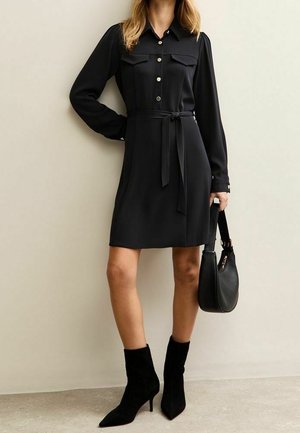 Femme portant une robe chemise noire à manches longues ceinturée avec des boutons dorés, des bottines noires, et tenant un sac à main noir.