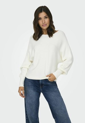 Pull en maille côtelée blanche avec un col large et des manches longues légèrement bouffantes, associé à un jean bleu foncé à jambes larges.