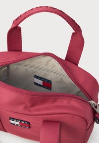 Kinyitott kis piros nylon táska textil fogantyúval, bézs belsővel és Tommy Jeans logóval ellátott foltokkal az elülső részen és a belső zsebében.