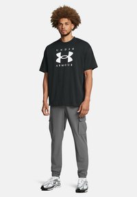 Schwarzes Baumwoll-T-Shirt mit weißem "UNDER ARMOUR"-Logo, kombiniert mit grauen Cargo-Hosen mit Seitentaschen und elastischen Bündchen. Schwarz-weiße Sneakers.