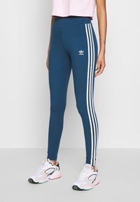 Legíny Adidas v odstínu teal s bílými bočními pruhy, design s vysokým pasem a elastickým materiálem. Nosené s bílými a černými sportovními botami.