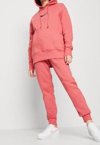 Person klädd i korallrosa Nike-hoodie och matchande sweatpants med vita Nike-sneakers, stående mot en enkel ljus bakgrund.