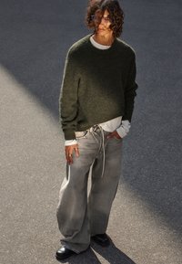 Olivgrüner, übergroßer Pullover, übereinander getragen mit einem weißen Langarmhemd. Hellgraue, weit geschnittene Jeans mit Kordelzug, schwarze klobige Schuhe.