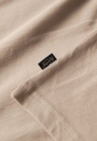 T-shirt en coton beige avec un col replié, présentant une étiquette noire avec le texte doré "Superdry" près de l'ourlet et des détails de couture visibles.