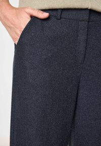 Main dans la poche d'un pantalon en laine gris foncé associé à un pull en tricot beige.