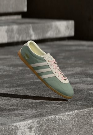 Baskets Adidas Gazelle vertes et blanches avec des lacets roses flottant au-dessus de marches en béton sous une lumière naturelle.