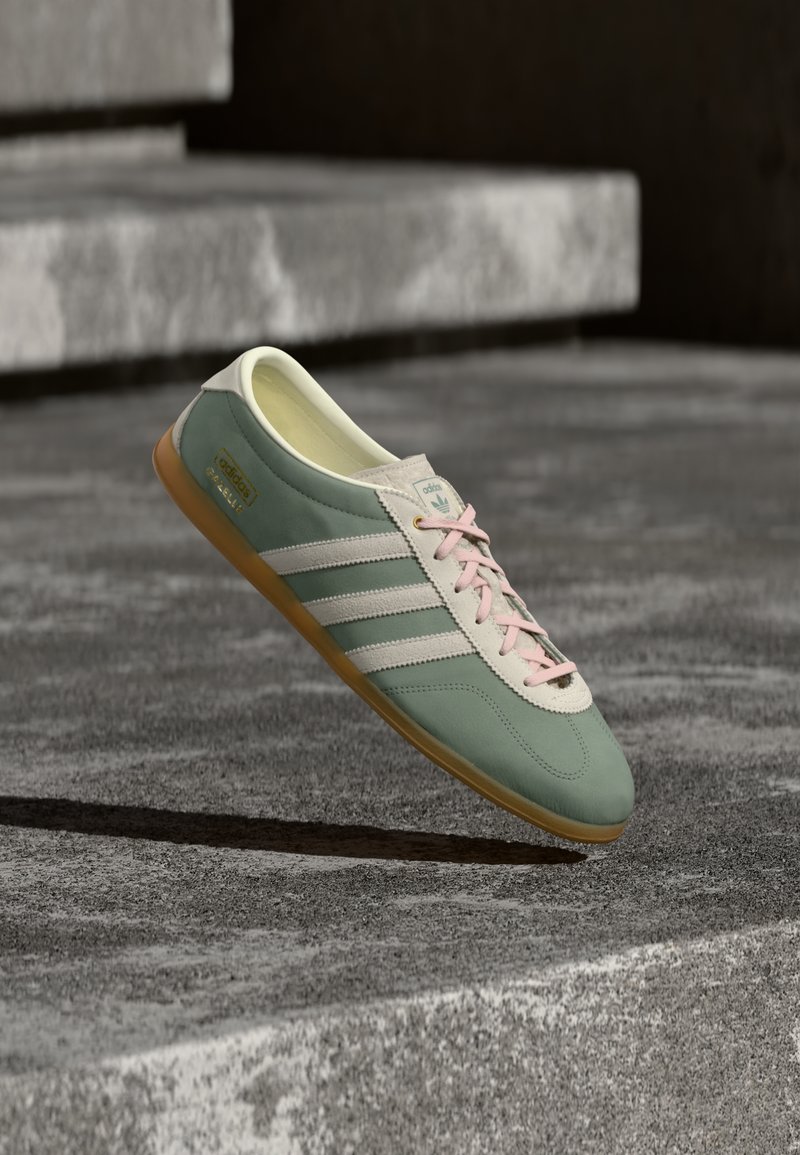 Πράσινο και λευκό αθλητικό παπούτσι Adidas Gazelle με ροζ κορδόνια, αιωρούμενο πάνω από σκάλες από σκυρόδεμα με φυσικό φωτισμό.