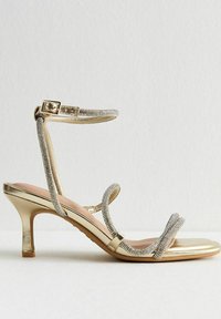 Goldene High Heels Sandalen mit dünnen, glitzernden Riemen über den Zehen und dem Knöchel, ausgestattet mit einem kleinen Schnallenverschluss und einem Absatz von mittlerer Höhe.