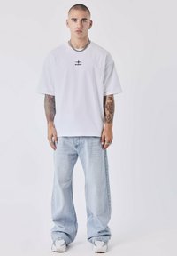 Witte t-shirt met korte mouwen en een zwart grafisch logo, gecombineerd met lichtblauwe baggy jeans en witte sneakers met grijze accenten.
