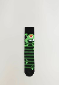 Chaussette rayée verte et noire présentant un design de monstre ludique avec un grand œil et un sourire décharné. Fabriquée en matériau tricoté doux.