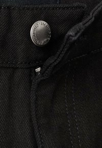 Material de denim negru cu cusături vizibile și un buton metalic etichetat "Înființat 1984". Vedere din apropiere a zonei taliei.