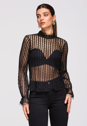 Mujer con top negro transparente de encaje de manga larga y cuello alto, con un bralette negro debajo, combinado con pantalones negros sobre un fondo liso.